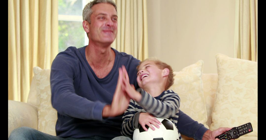 Father and Son Laughing on Couch with Soccer Ball and TV Remote - Free Images, Stock Photos and Pictures on Pikwizard.com