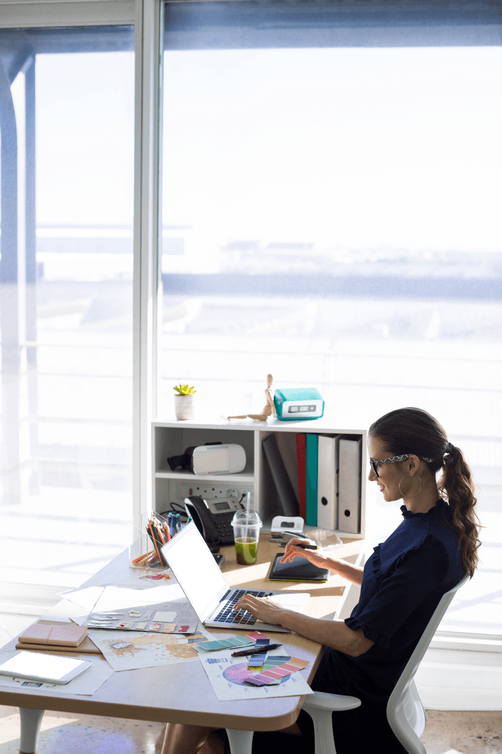 Businesswoman Working on Laptop at Desk in Bright Office with Transparent Window
