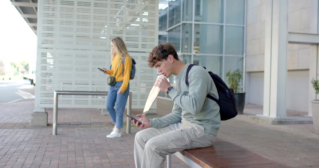 College student checking phone while opening takeout bag on campus bench with backpack