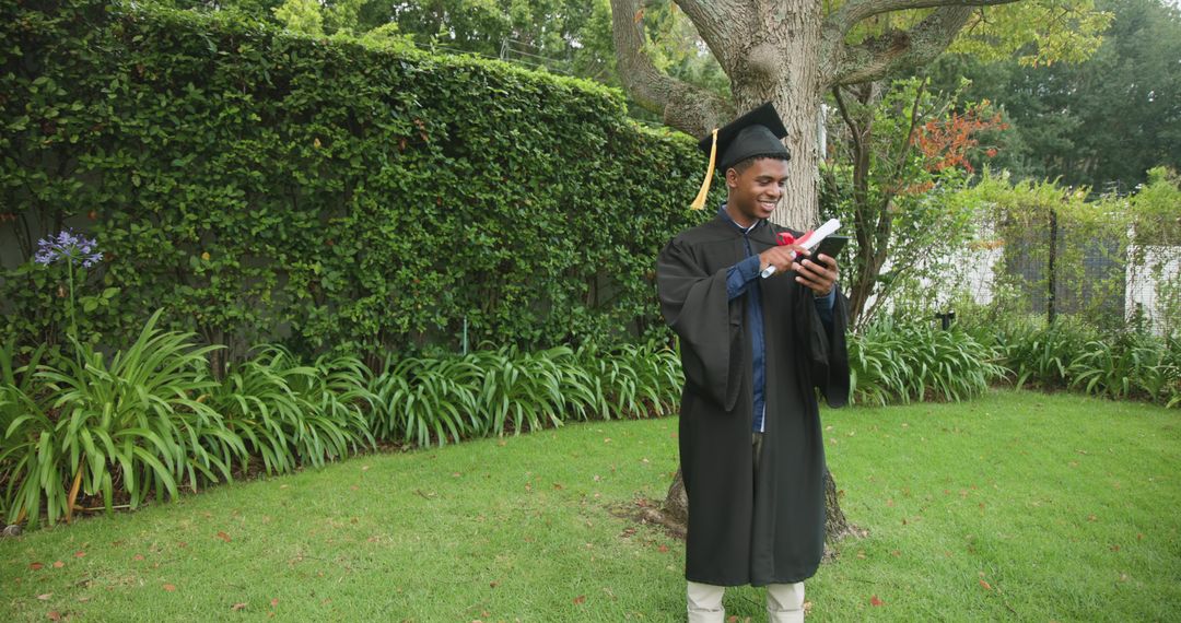 Graduate wearing cap checking smartphone while holding diploma on green lawn under tree