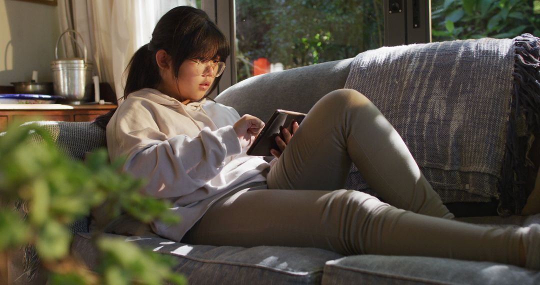 Asian girl relaxing on couch using digital tablet in sunlit living room - Free Images, Stock Photos and Pictures on Pikwizard.com