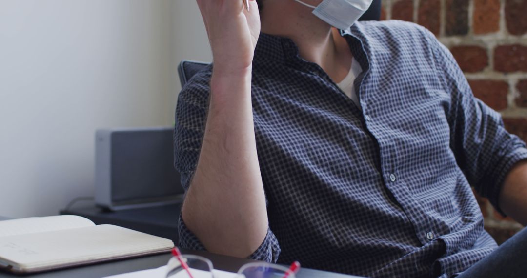 Man Wearing Face Mask Working in Modern Office with Notebook and Glasses on Desk - Free Images, Stock Photos and Pictures on Pikwizard.com