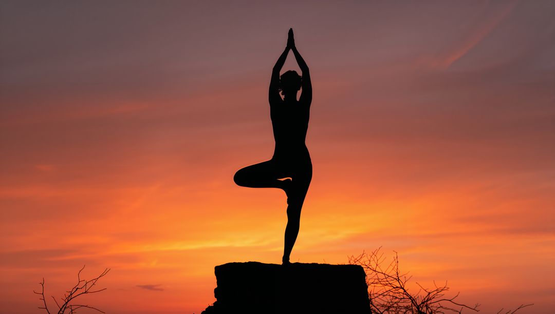 Silhouetted woman practicing tree pose on cliff at vibrant orange and purple sunset