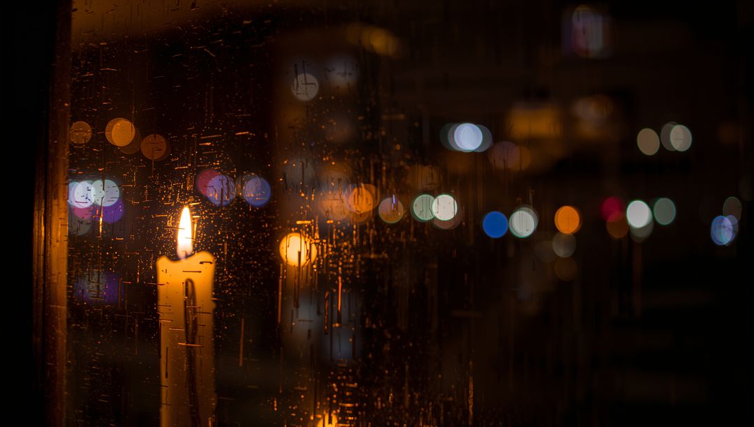 Glowing pillar candle melting wax reflecting flame on rain-speckled glass with city bokeh