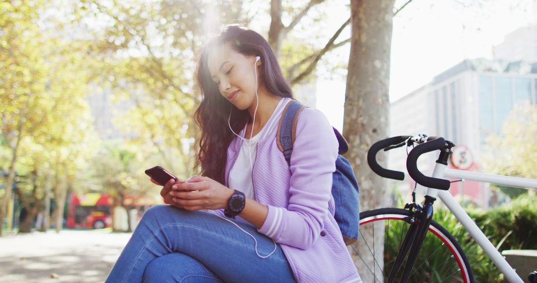 Asian woman wearing earphones using smartphone sitting in the park - Free Images, Stock Photos and Pictures on Pikwizard.com