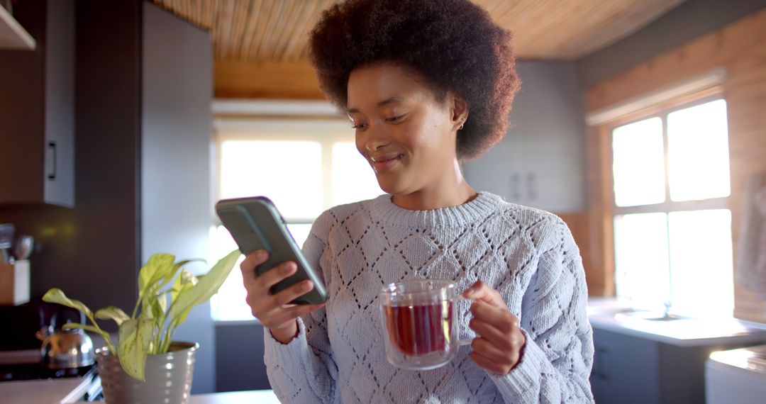 Happy african american woman with afro holding cup of tea and using smartphone in sunny kitchen - Free Images, Stock Photos and Pictures on Pikwizard.com