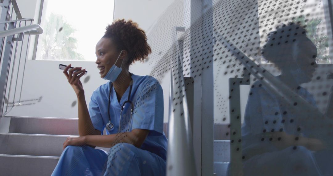 Nurse speaking into smartphone while resting on hospital stairs with mask and stethoscope