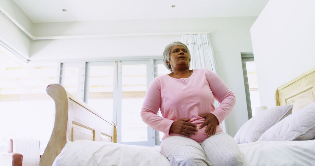 Elderly Woman Experiencing Discomfort Sitting on Bed Holding Stomach - Free Images, Stock Photos and Pictures on Pikwizard.com