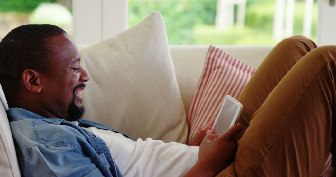 Relaxed Man Laughing at Digital Device on Sofa at Home - Free Images, Stock Photos and Pictures on Pikwizard.com