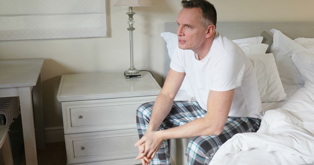 Middle-aged man reflecting while sitting on bed in plaid pajama pants - Free Images, Stock Photos and Pictures on Pikwizard.com