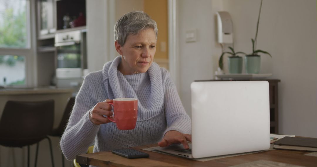 Senior woman working on laptop while drinking coffee in cozy kitchen - Free Images, Stock Photos and Pictures on Pikwizard.com