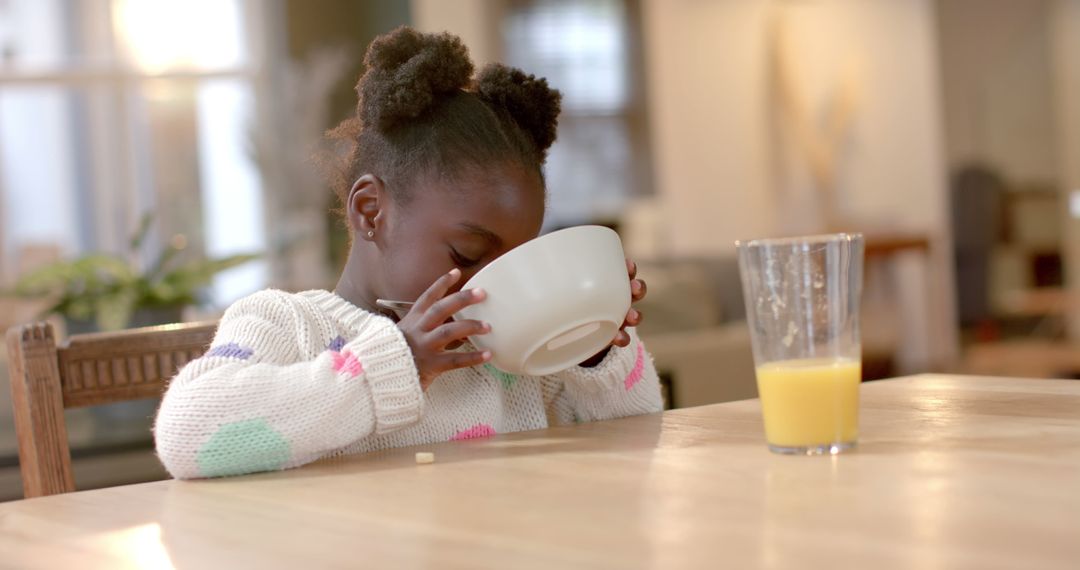 Young Girl Drinking from Bowl Sitting at Table with Glass of Orange Juice - Free Images, Stock Photos and Pictures on Pikwizard.com