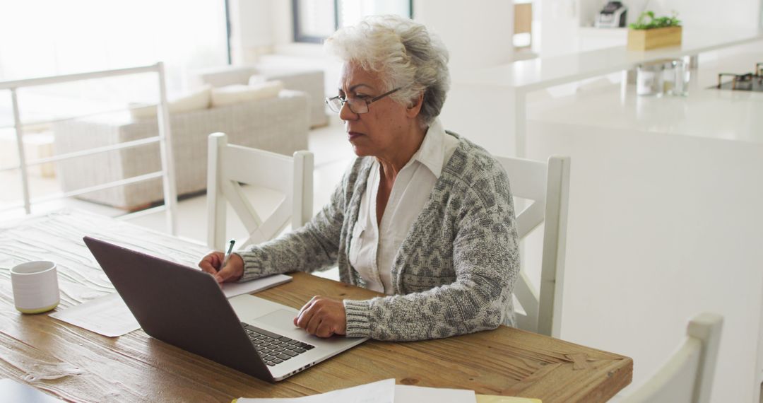 Elderly Woman Using Laptop While Taking Notes in Bright Modern Home - Free Images, Stock Photos and Pictures on Pikwizard.com