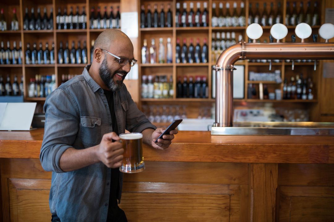 Man Enjoying Beer and Using Smartphone at Wooden Bar Counter