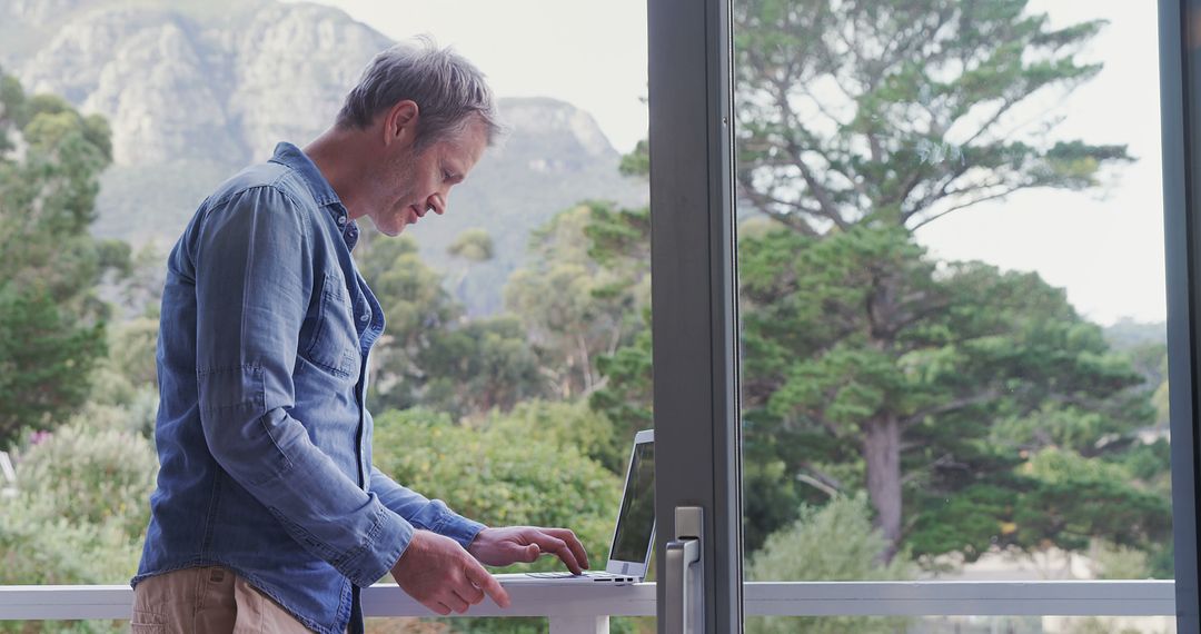 Middle-aged man working on laptop by window with scenic view - Free Images, Stock Photos and Pictures on Pikwizard.com