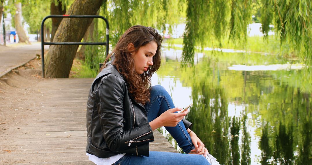 Young Woman Sitting by Tranquil Lake Texting on Smartphone - Free Images, Stock Photos and Pictures on Pikwizard.com