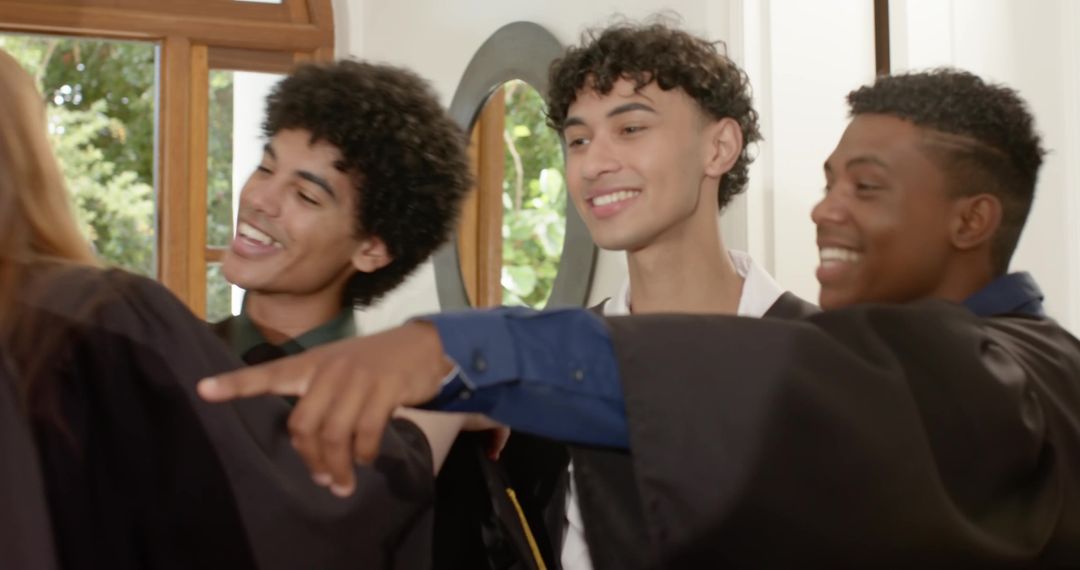 Smiling diverse graduates posing in black gowns, pointing and celebrating achievement by sunlit mirr