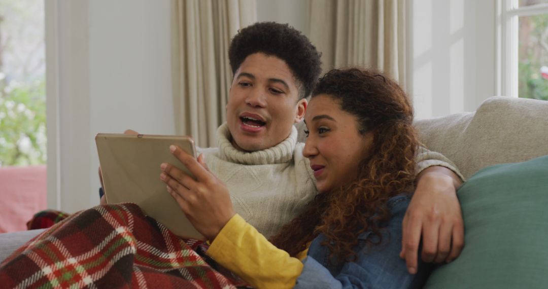 Image of happy biracial couple siting on sofa under blanket and using tablet - Free Images, Stock Photos and Pictures on Pikwizard.com