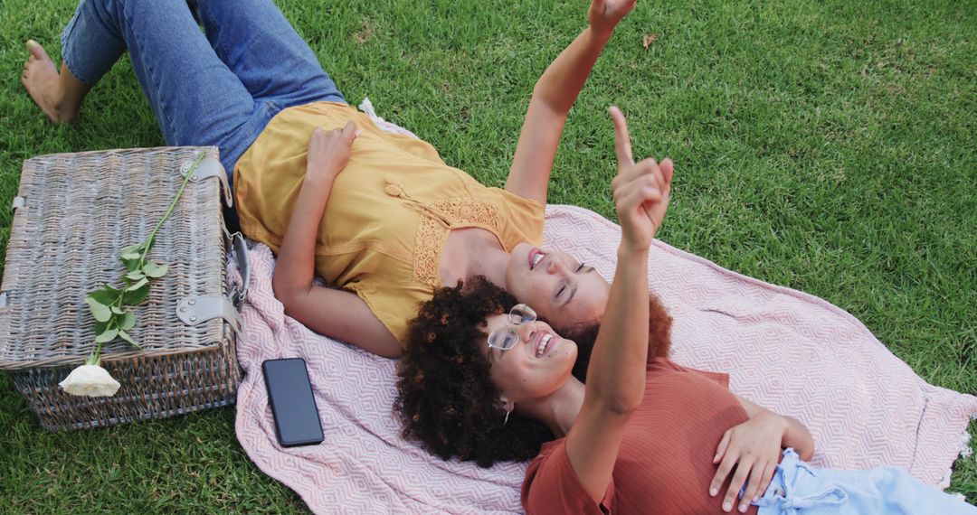 Two women lying on picnic blanket pointing at sky with wicker basket and smartphone