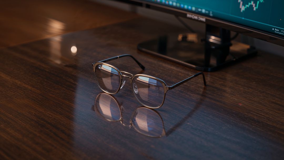 Eyeglasses on Office Desk Next to Monitor with Financial Graphs