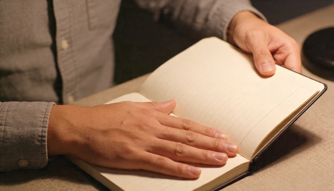 Man Holding Open Lined Notebook on Desk While Preparing to Write Under Warm Lamp Light