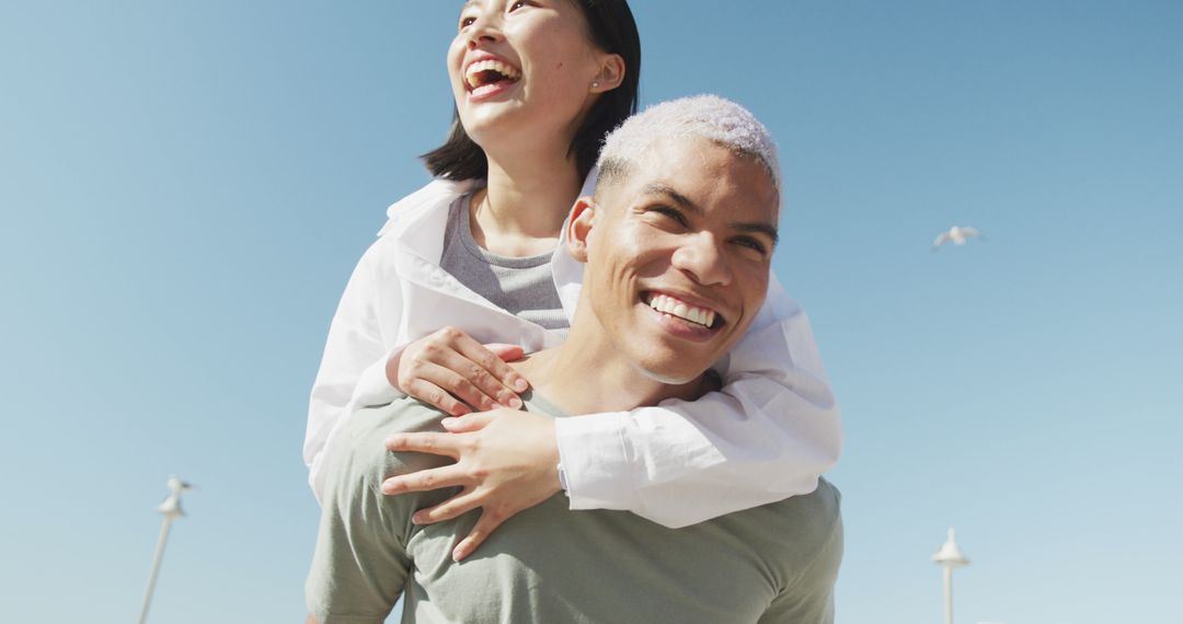 Happy Multiethnic Couple Laughing Piggyback at Beach on Sunny Day - Free Images, Stock Photos and Pictures on Pikwizard.com