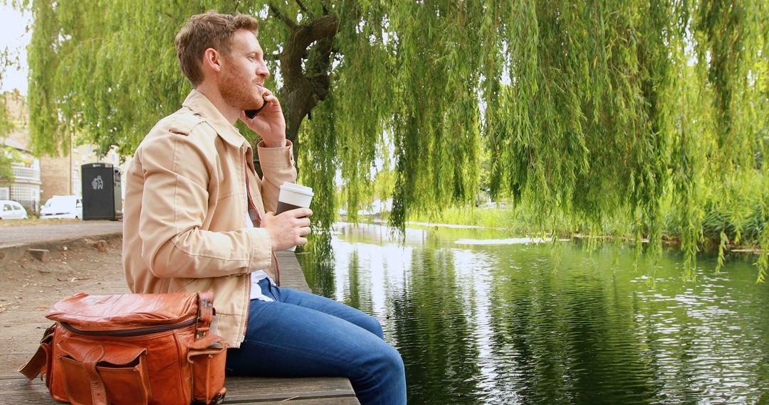 Young man talking on phone holding coffee by lake under willow tree - Free Images, Stock Photos and Pictures on Pikwizard.com