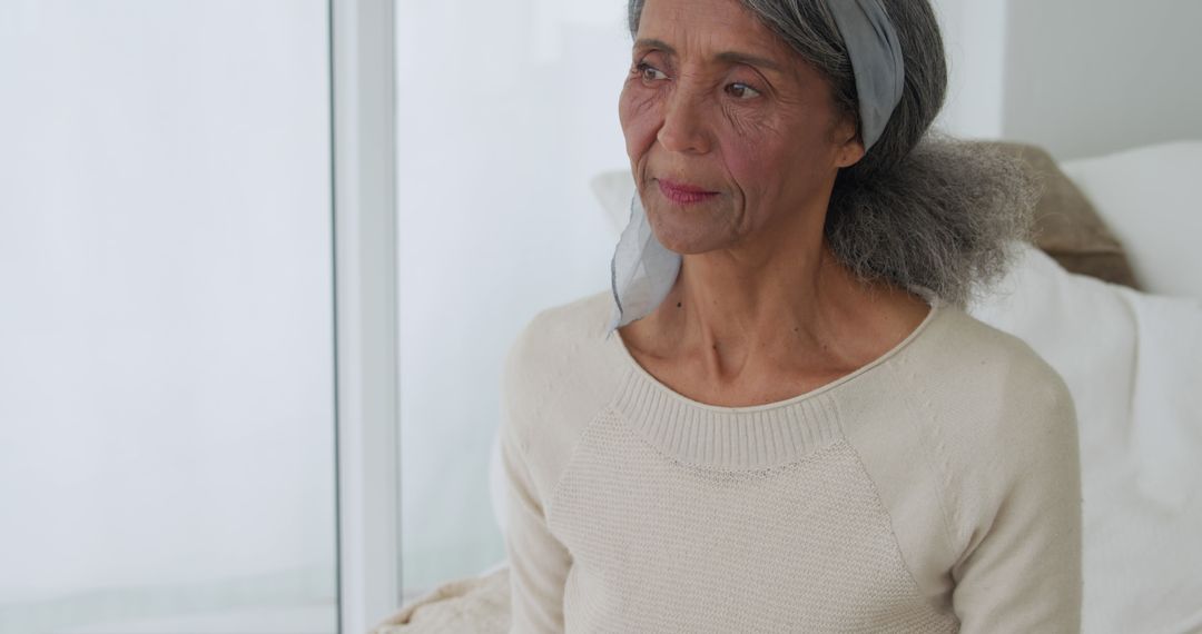 Pensive Elderly Woman Sitting By Window Wearing Grey Headband - Free Images, Stock Photos and Pictures on Pikwizard.com