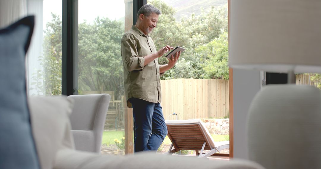 Middle-aged man using digital tablet in modern living room with garden view - Free Images, Stock Photos and Pictures on Pikwizard.com