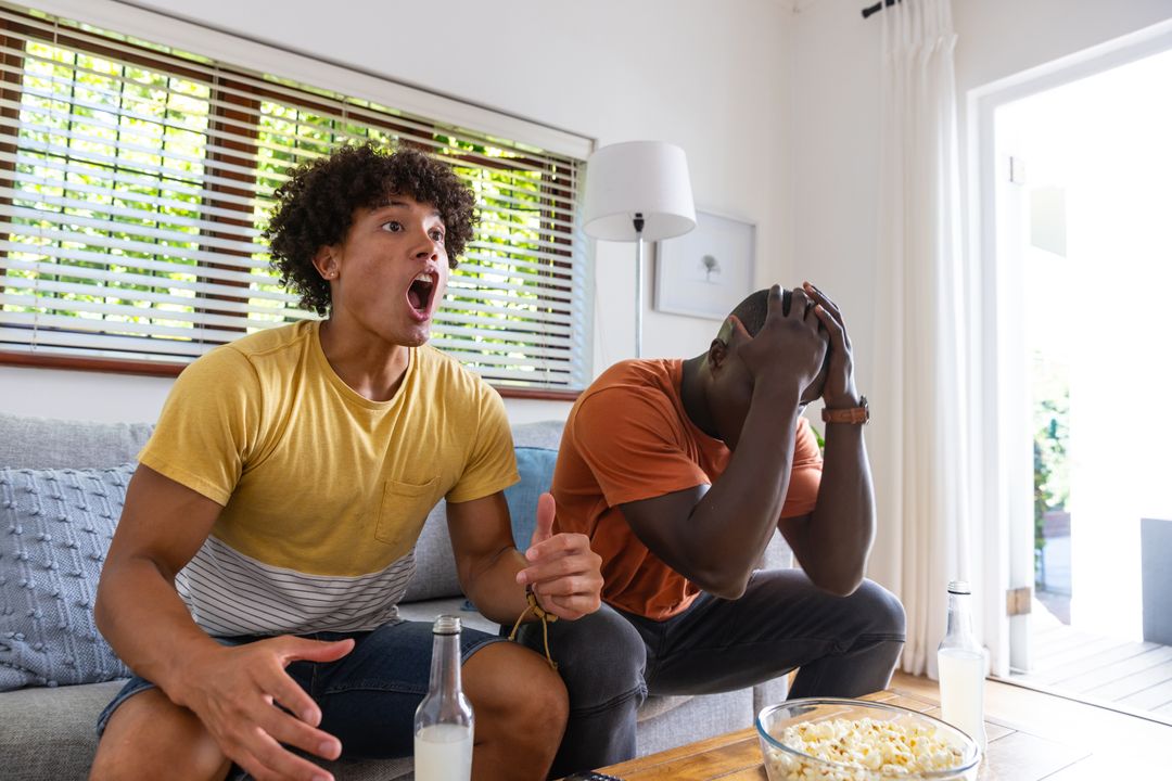 Two diverse male friends reacting while watching tense football match on couch with snacks