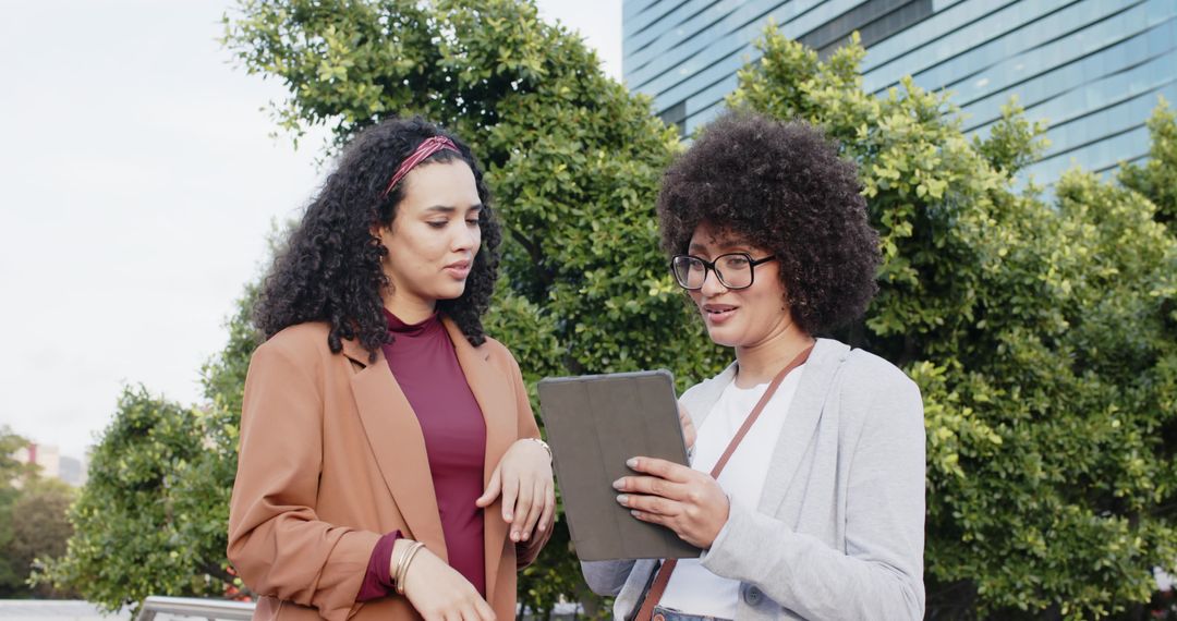 Female colleagues reviewing tablet in modern urban courtyard, collaborative business moment