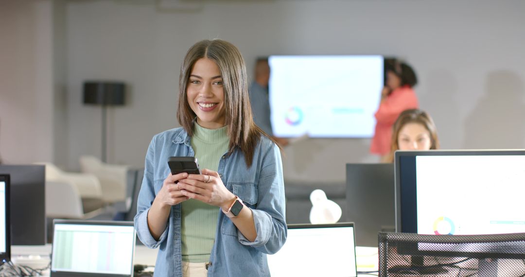 Young professional woman smiling while using smartphone in modern open office with teamwork