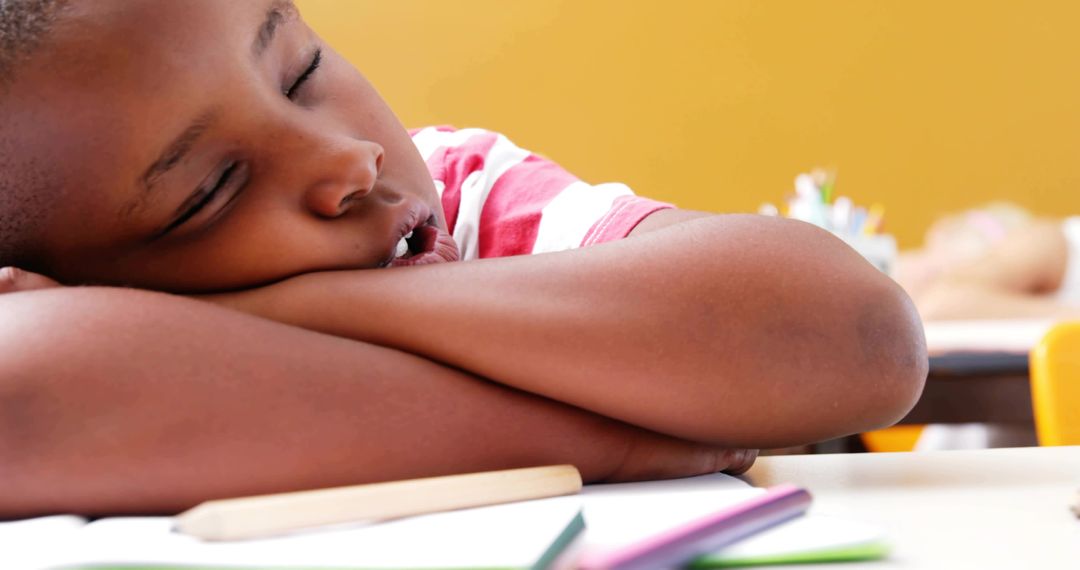 Sleepy Schoolchild Resting on Desk with Colored Pencils and Yellow Classroom Background