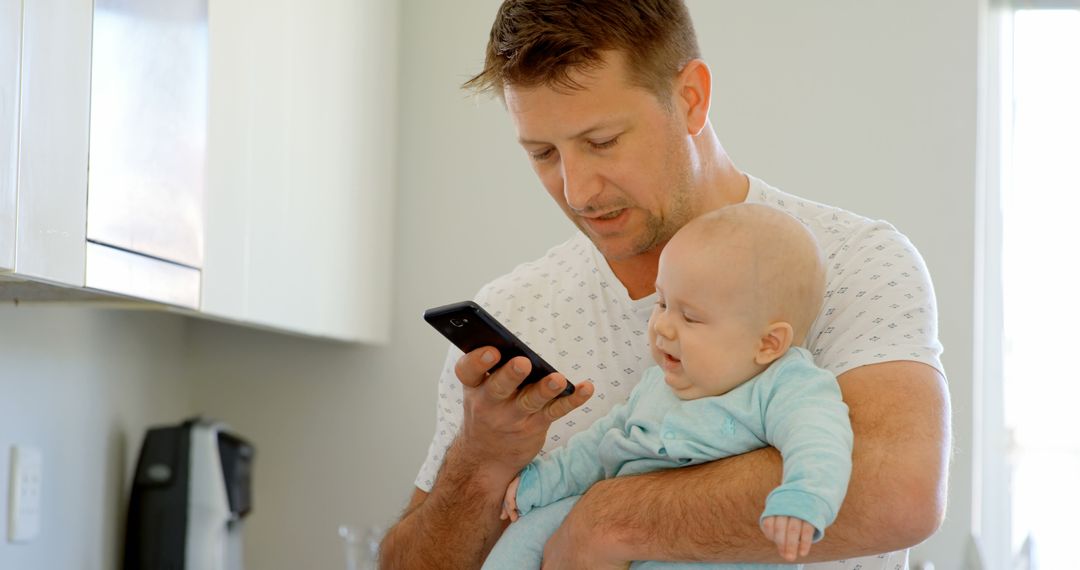 Father Holding Baby While Using Smartphone in Kitchen - Free Images, Stock Photos and Pictures on Pikwizard.com