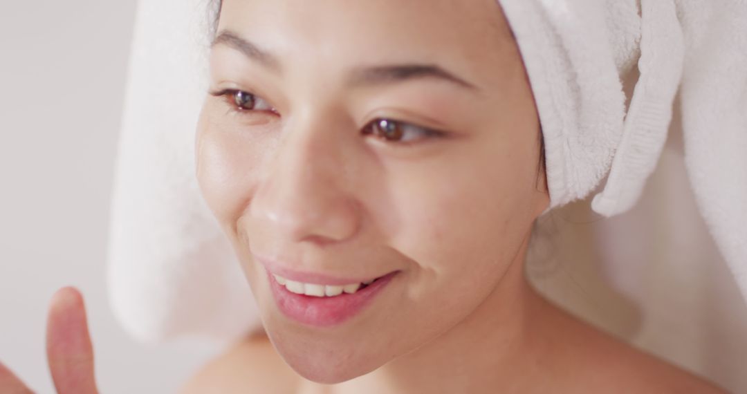 Image of portrait of smiling biracial woman with towel on hair applying face serum in bathroom - Free Images, Stock Photos and Pictures on Pikwizard.com