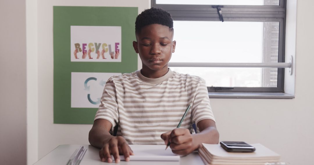 Focused African American Teen Writing in Classroom with Books and Smartphone
