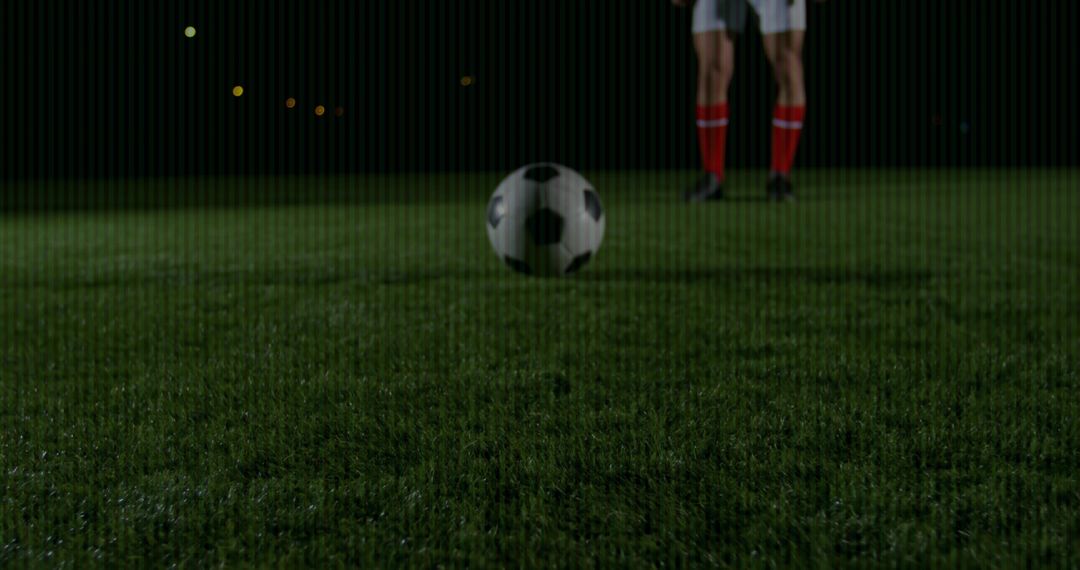 Hovering soccer ball over wet pitch at night while player standing in red knee-high socks