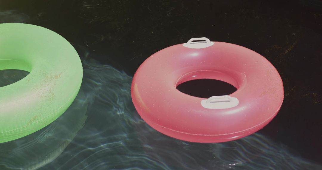 Sunlit pink and green inflatable rings floating on dark pool water with reflective ripples