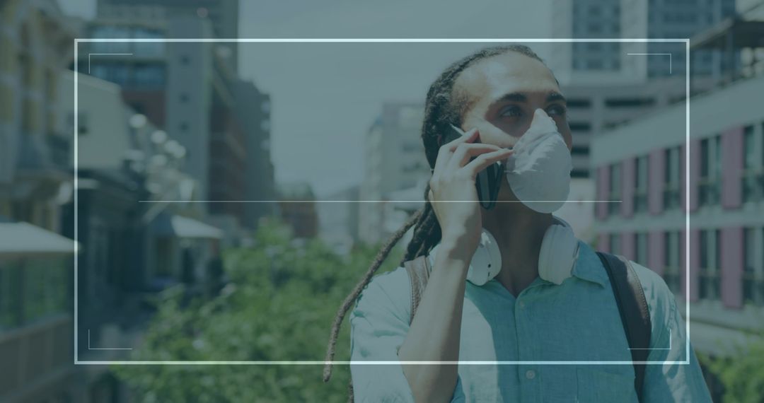 Young man wearing mask talking on phone while walking on urban elevated walkway with headphones and 