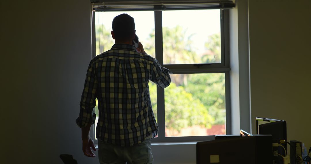 Man Conversing On Phone By Window In Office With Greenery Outside - Free Images, Stock Photos and Pictures on Pikwizard.com