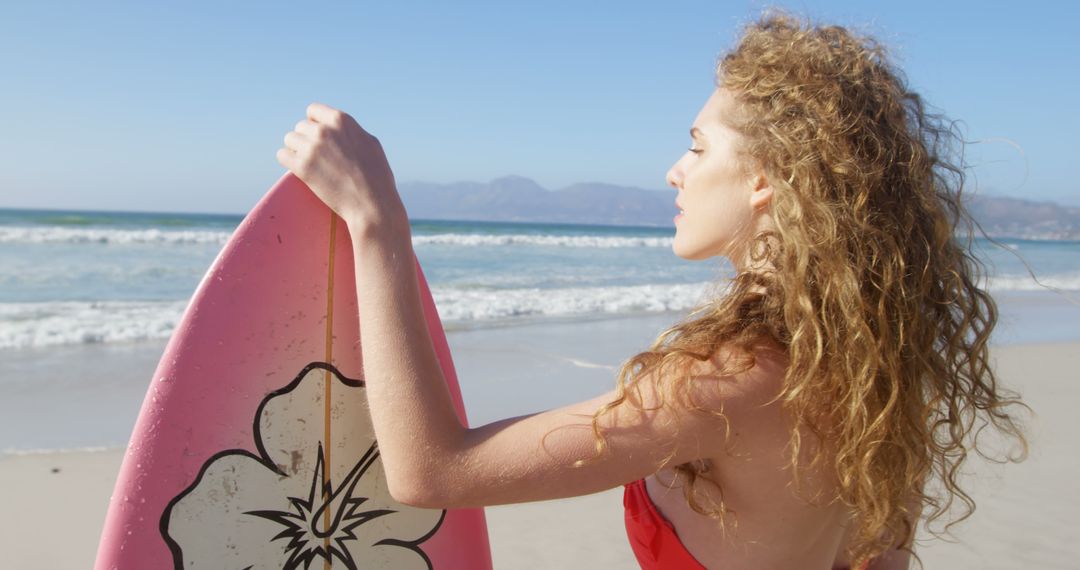 A young Caucasian woman in a red swimsuit holds a pink surfboard on a sunny beach, with copy space - Free Images, Stock Photos and Pictures on Pikwizard.com