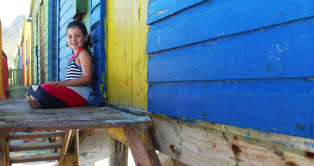 Joyful Girl Sitting by Colorful Beach Huts on a Sunny Day - Free Images, Stock Photos and Pictures on Pikwizard.com