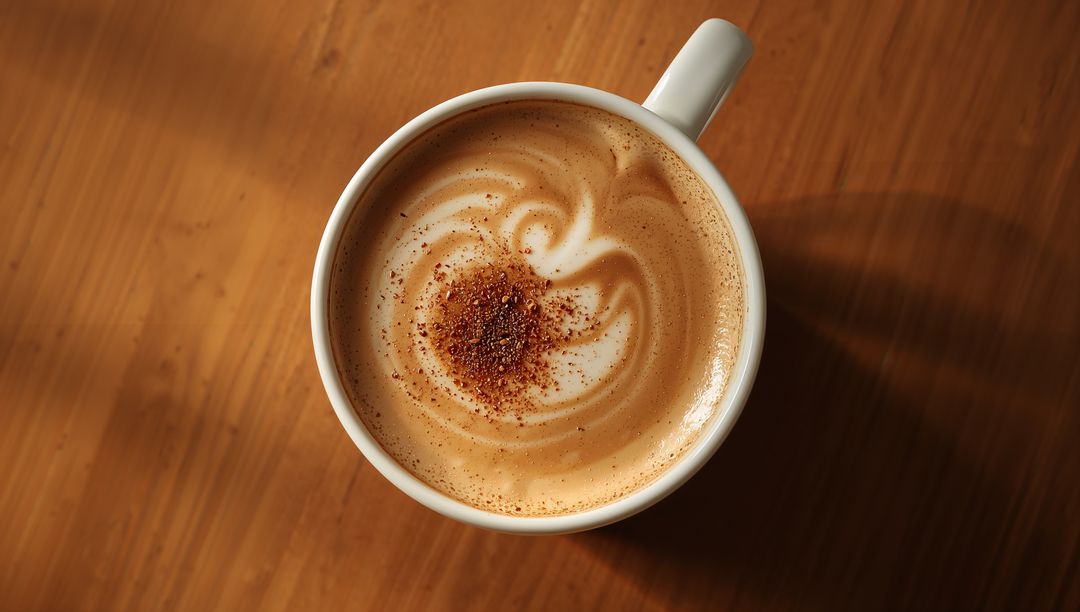 Creamy latte with cinnamon dust in white mug on warm wood countertop, top-down flatlay