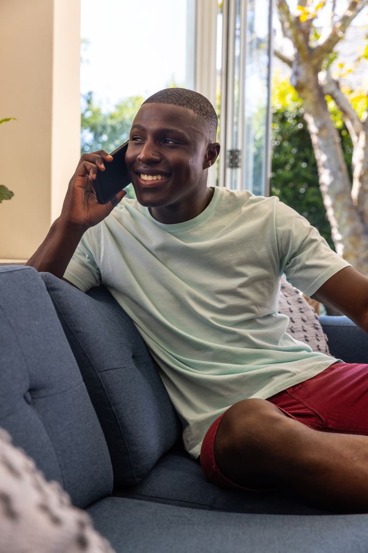 African American man relaxing on sofa smiling while talking on smartphone near open doors