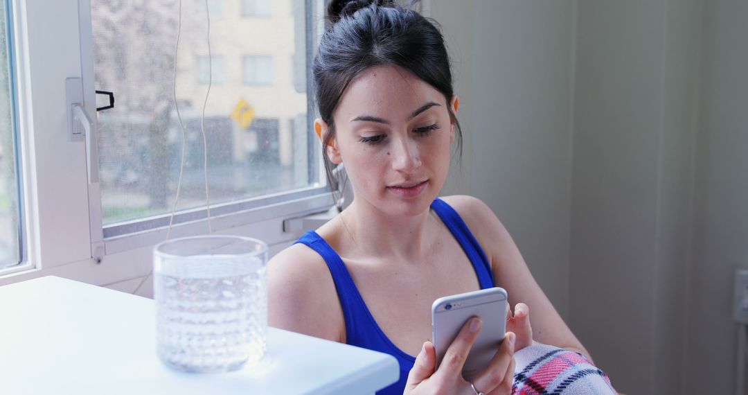 Young Woman Using Smartphone Next to Window with Glass of Water - Free Images, Stock Photos and Pictures on Pikwizard.com