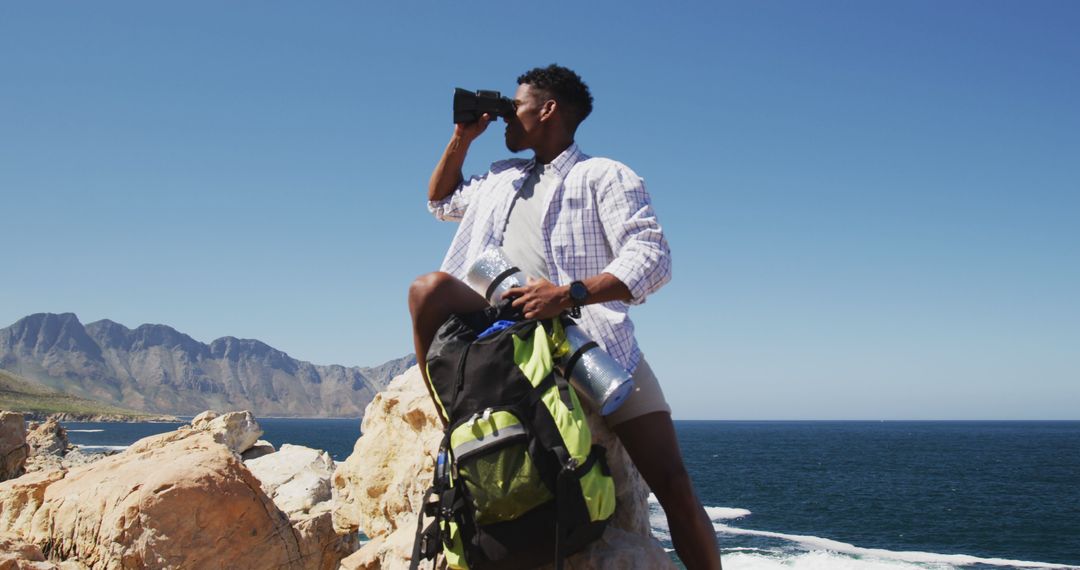 African american man hiking using binocilars sitting on rock by the coast - Free Images, Stock Photos and Pictures on Pikwizard.com