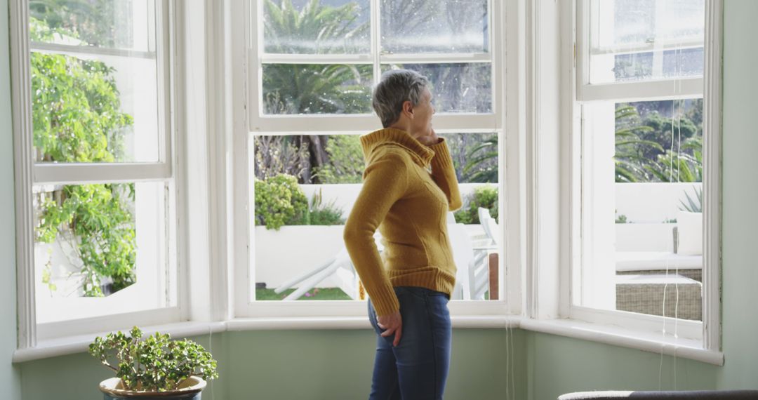 Mature Woman in Sweater Looking Out Bay Window at Green Garden - Free Images, Stock Photos and Pictures on Pikwizard.com