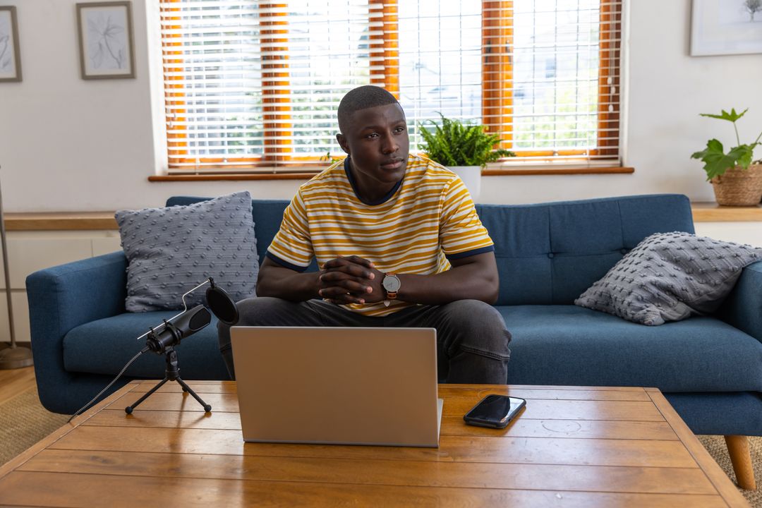 African American man relaxing on couch using laptop and podcast microphone at home