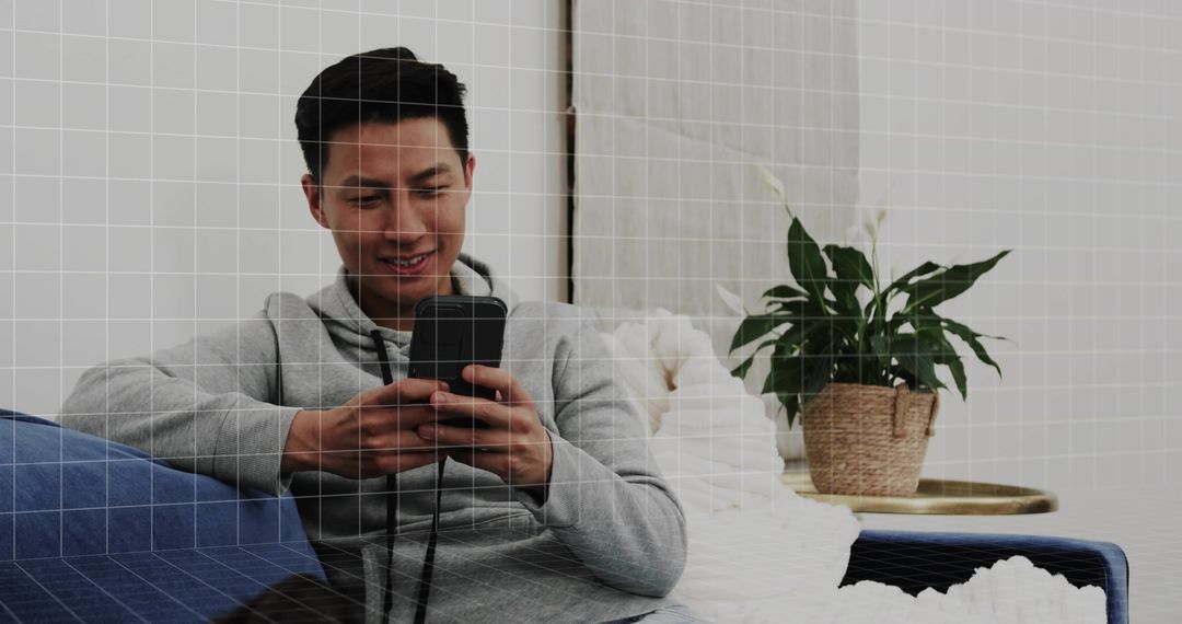 Young man relaxing on blue sofa scrolling smartphone with houseplant and cozy linens