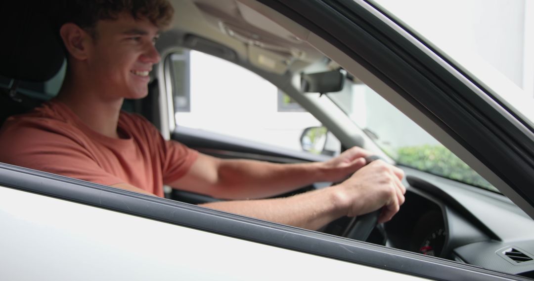 Young man smiling while driving white car through open window wearing rust T-shirt